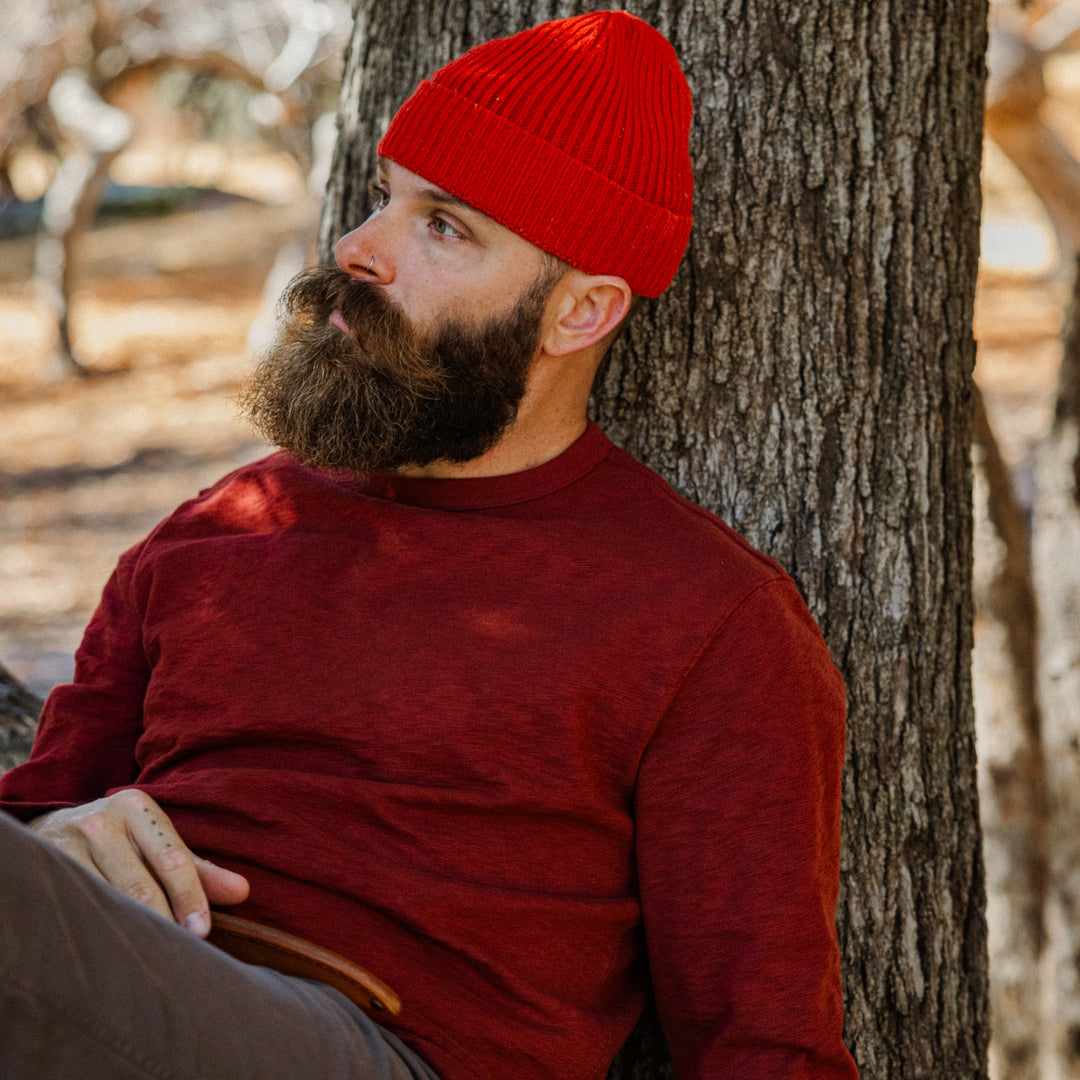 Man wearing a red beanie and Rugby Crew Crimson leaning against a tree in a forest setting