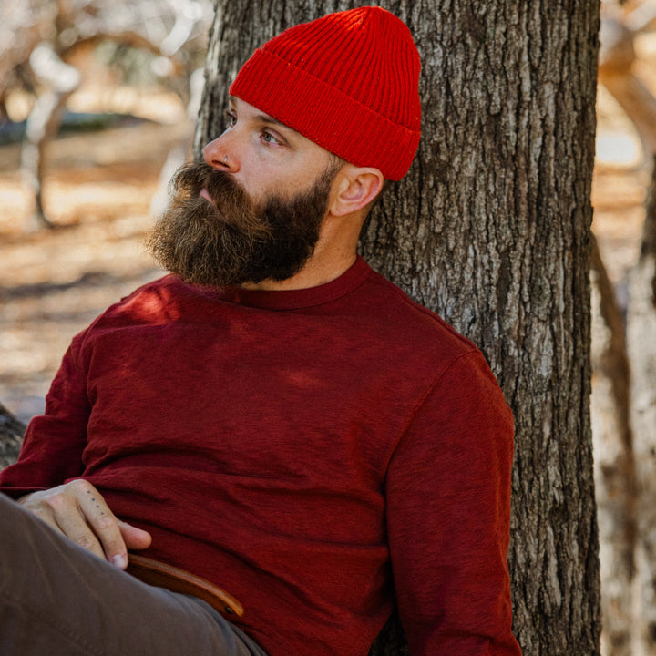 Man wearing a red beanie and Rugby Crew Crimson leaning against a tree in a forest setting