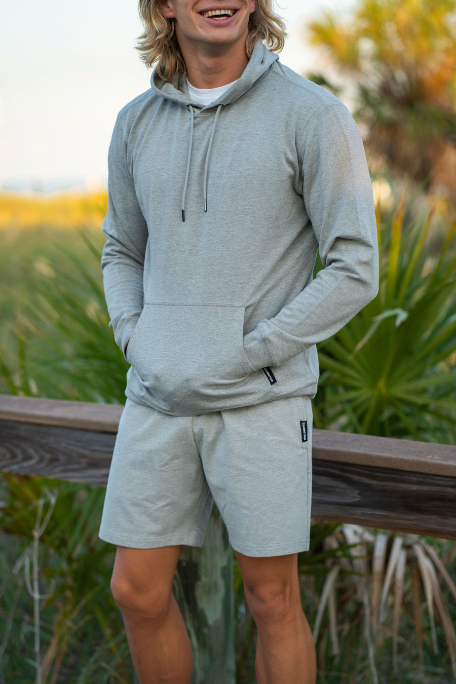 Man wearing Lounge Hoodie in Heather Grey and Lounge Shorts in Heather Grey standing outdoors with greenery in the background