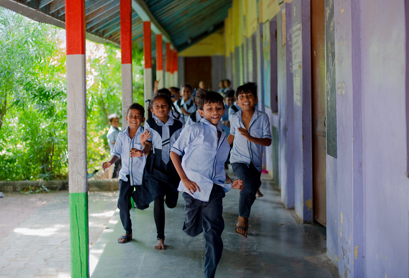 A group of young school children running happily along a covered outdoor corridor with colorful pillars and walls.