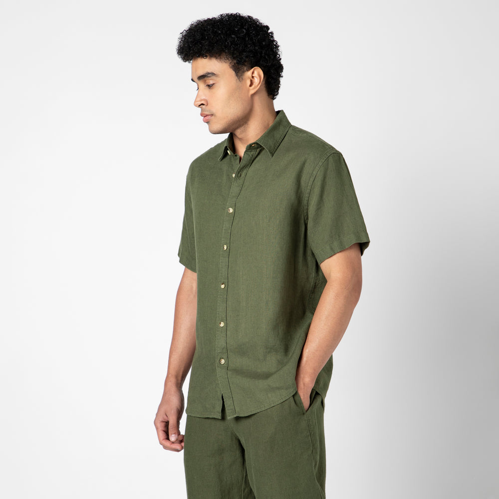 A man with short curly hair models the Linen Short Sleeve Shirt in olive green and matching pants against a plain white background, looking down to his left to showcase the breathable comfort of natural linen.