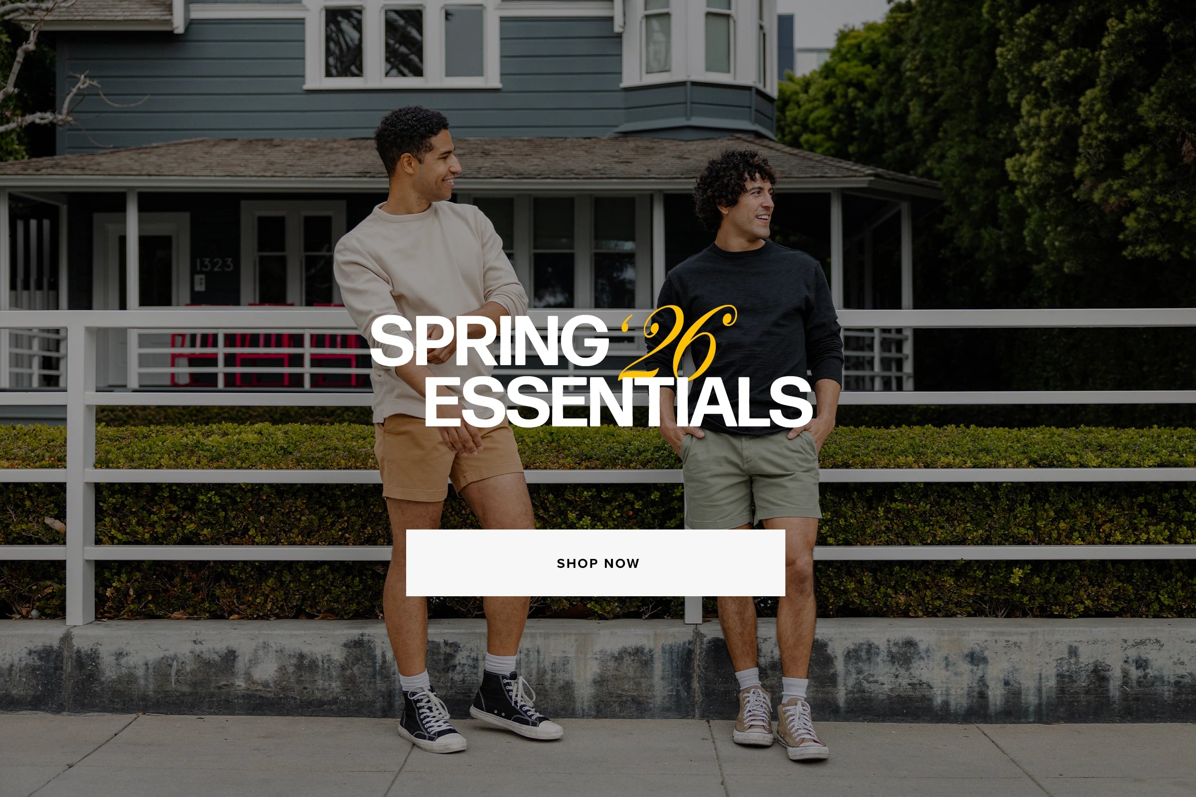 Two men standing in front of a house with 'Spring Essentials 2026' text overlay. Man on left wearing Thermal Knit Crew in Oat and Stretch Short in Camel and Man on right wearing Lounge Crewneck in Black and Stretch Short in Olive. Shop Spring Essentials Now.