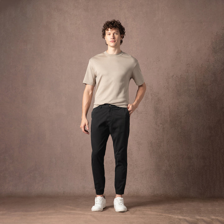 A person stands in the black Stretch Jogger with a beige short-sleeve tee and white sneakers against a warm brown studio backdrop, shown head to toe.