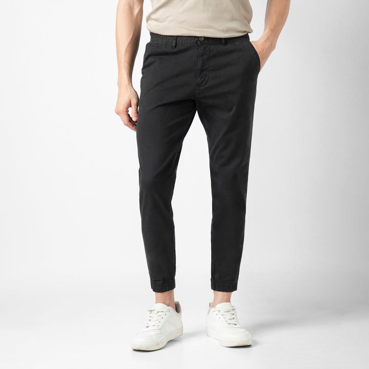 A person wears the black Stretch Jogger with an elastic waistband and tapered leg, paired with a beige short-sleeve tee and white sneakers, standing against a plain light grey background. The image is cropped at the shoulders.