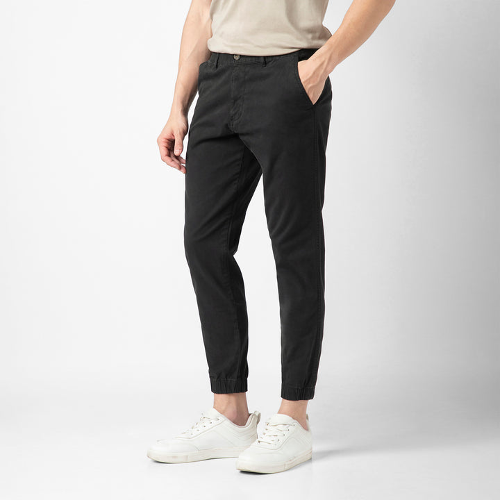 A person stands at an angle wearing the black Stretch Jogger with one hand tucked into the side pocket, along with a beige short-sleeve tee and white sneakers, against a plain light grey background. The image is cropped at the shoulders.
