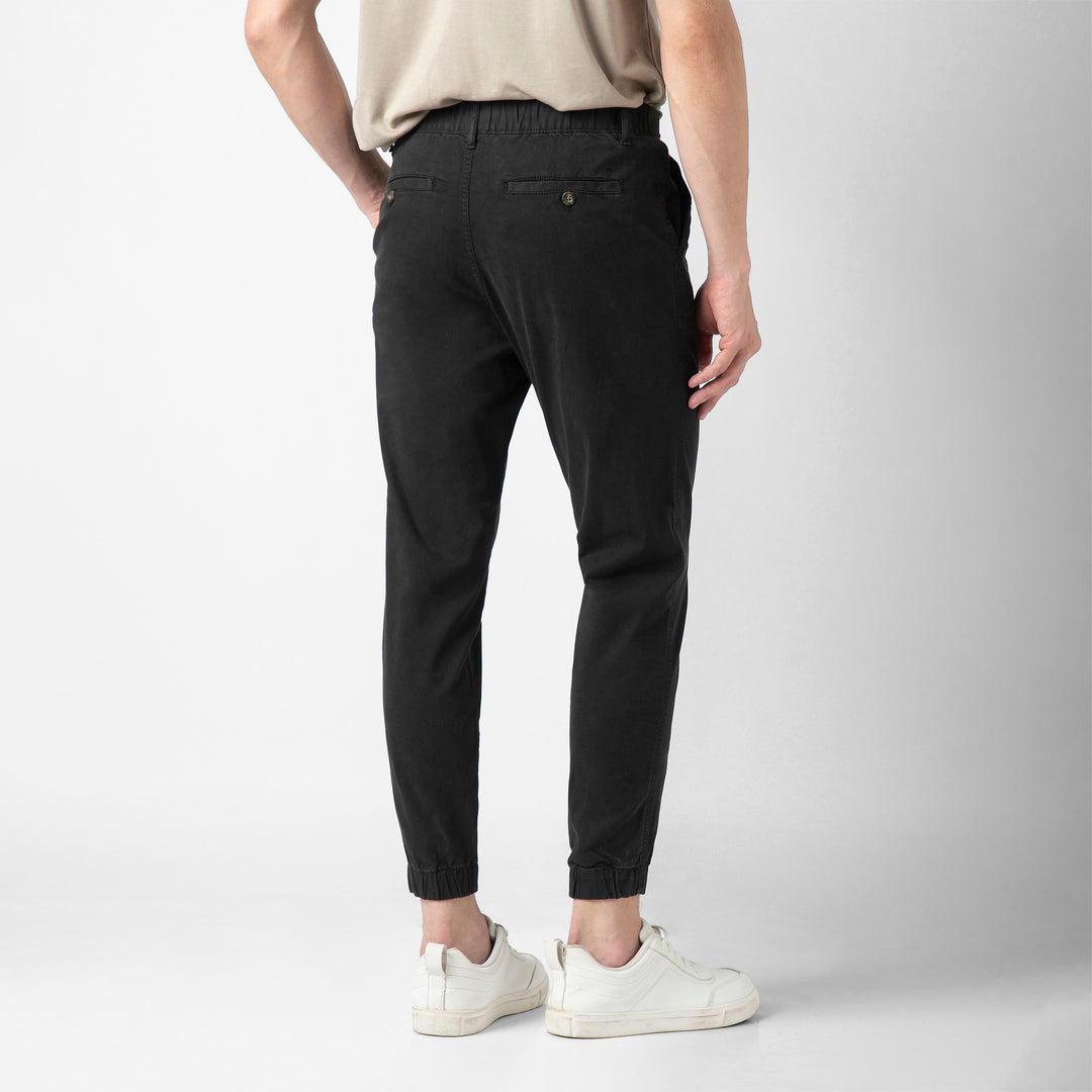 A person stands with their back to the camera wearing the black Stretch Jogger, showing the buttoned back pocket and elastic ankle cuffs, with a beige tee and white sneakers against a plain light grey background.