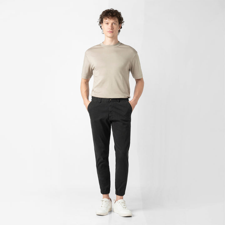 A person stands facing the camera in the black Stretch Jogger with a beige short-sleeve tee and white sneakers, shown head to toe against a plain light grey background.