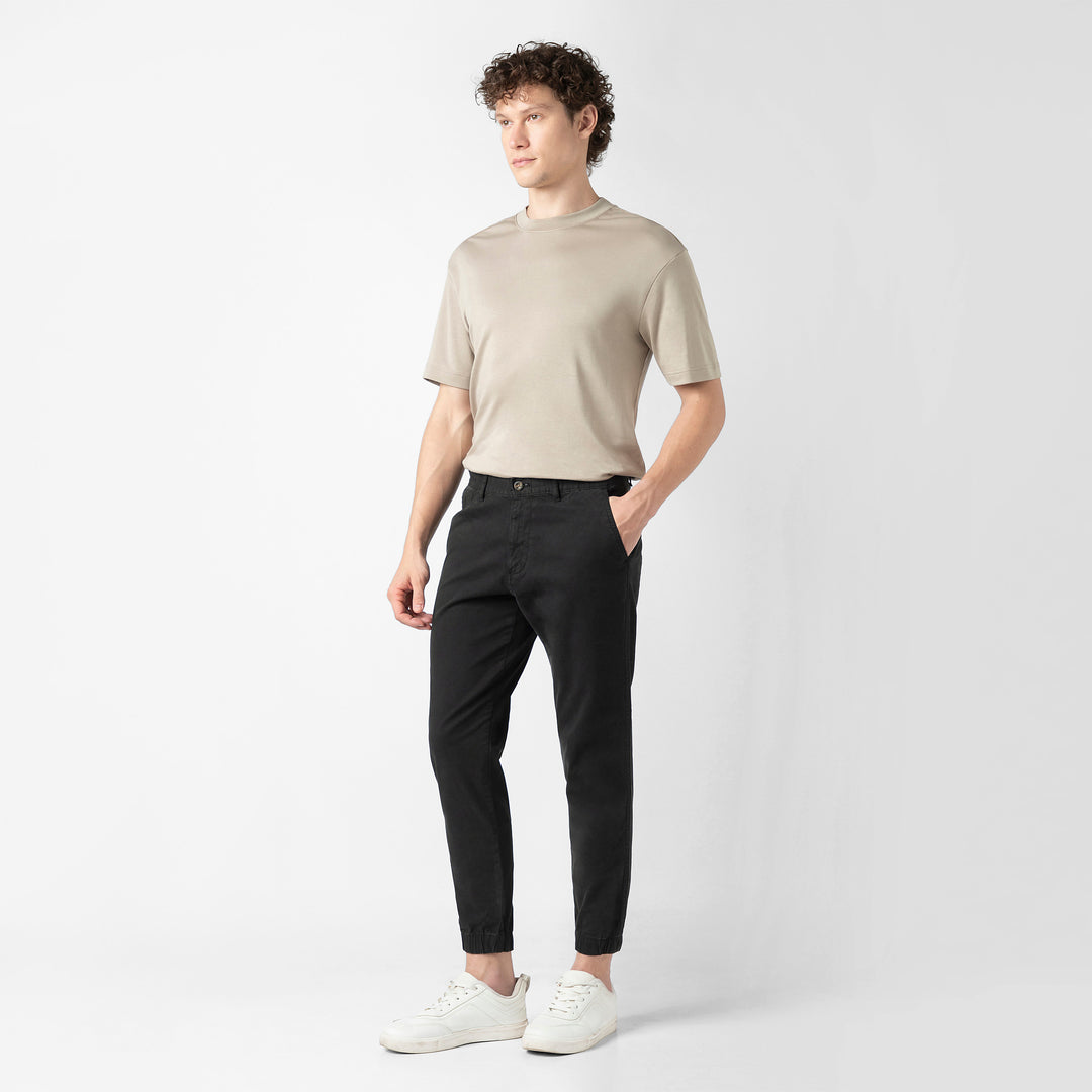 A person stands at a three-quarter angle in the black Stretch Jogger with one hand in the pocket, wearing a beige short-sleeve tee and white sneakers, shown head to toe against a plain light grey background.