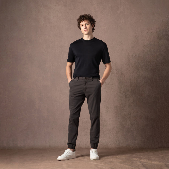 A person stands in the Coal Stretch Jogger with a black short-sleeve tee and white sneakers against a warm brown studio backdrop, shown head to toe.