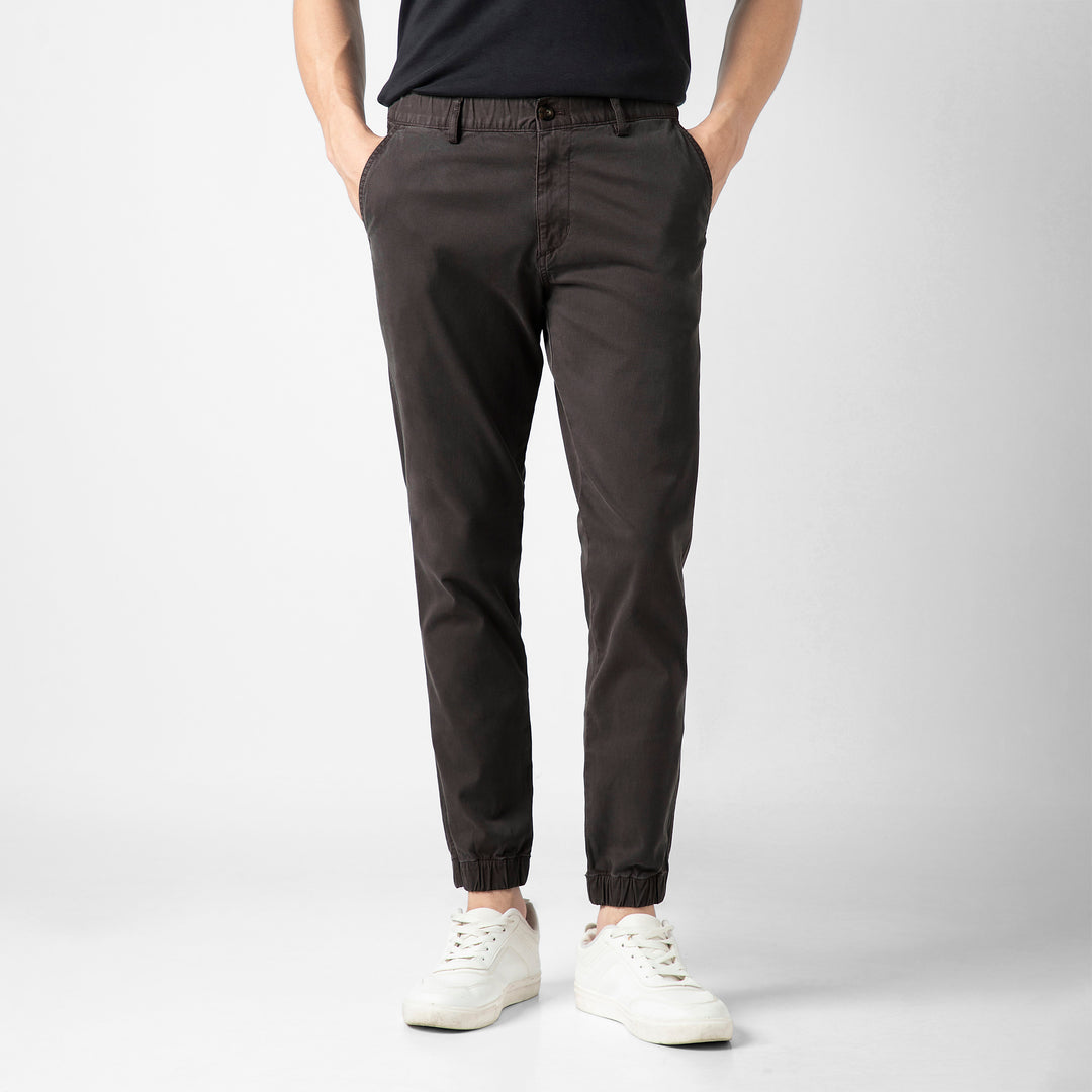 A person wears the Stretch Jogger in Coal with an elastic waistband, paired with a black short-sleeve tee and white sneakers, against a plain light grey background. The image is cropped at the shoulders.