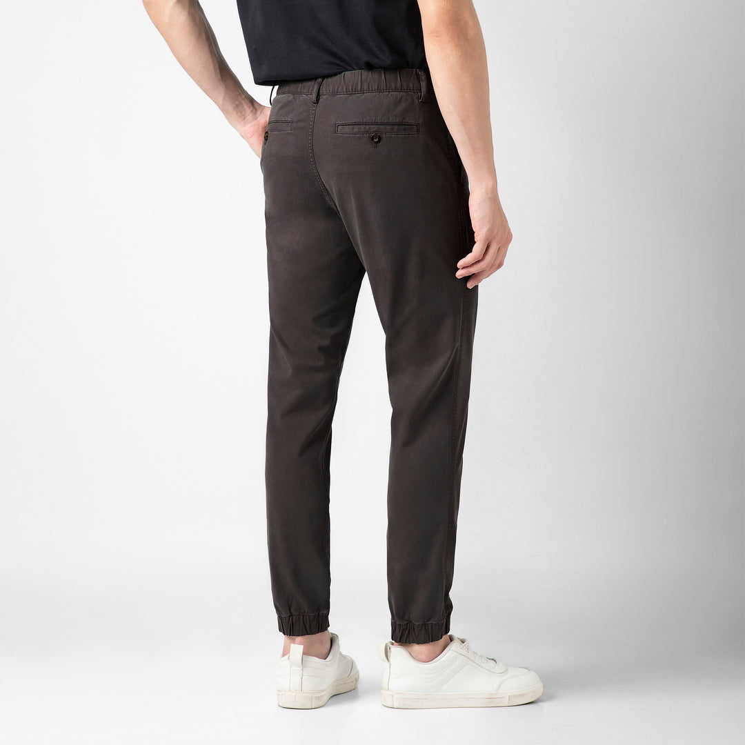 A person stands with their back to the camera in the Coal Stretch Jogger, showing the buttoned back pocket and elastic ankle cuffs, with a black tee and white sneakers.