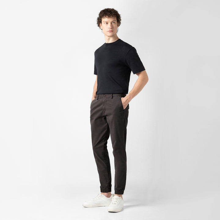 A person stands facing the camera in the Coal Stretch Jogger with a black short-sleeve tee and white sneakers, shown head to toe against a plain light grey background.