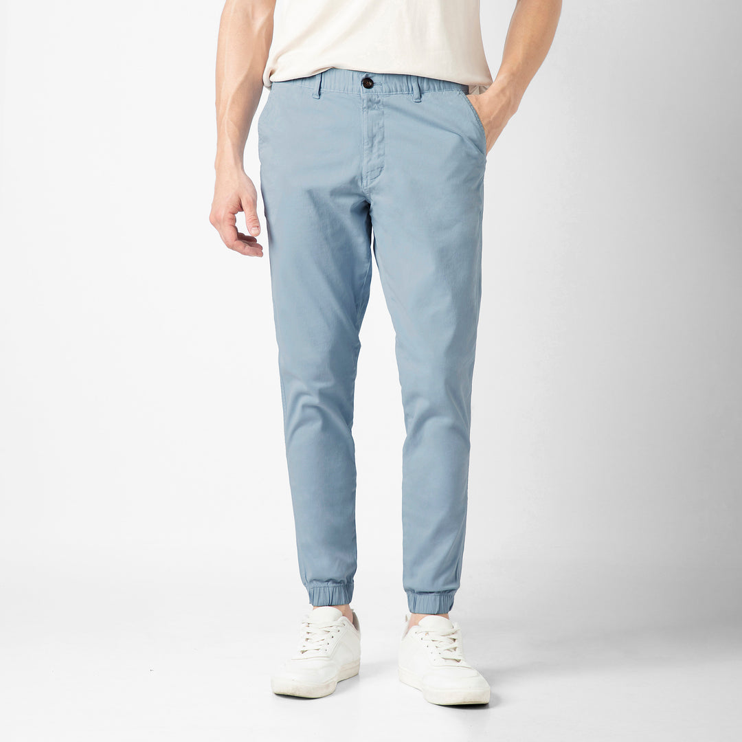 A person wears the Stretch Jogger in Coastal light blue with an elastic waistband, paired with a cream short-sleeve tee and white sneakers, against a plain light grey background. The image is cropped at the shoulders.
