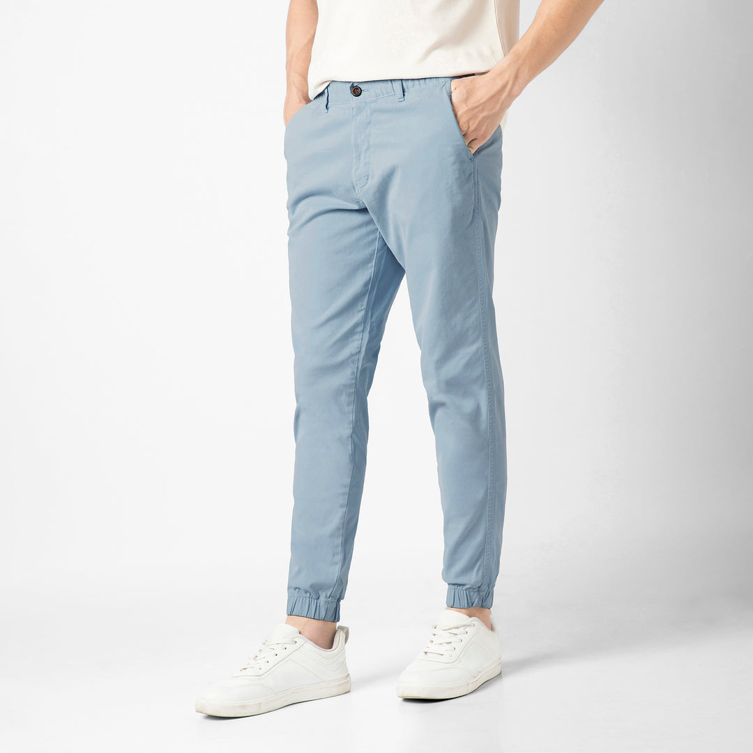 A person stands at an angle in the Coastal light blue Stretch Jogger with one hand tucked into the side pocket, wearing a cream tee and white sneakers against a plain light grey background.