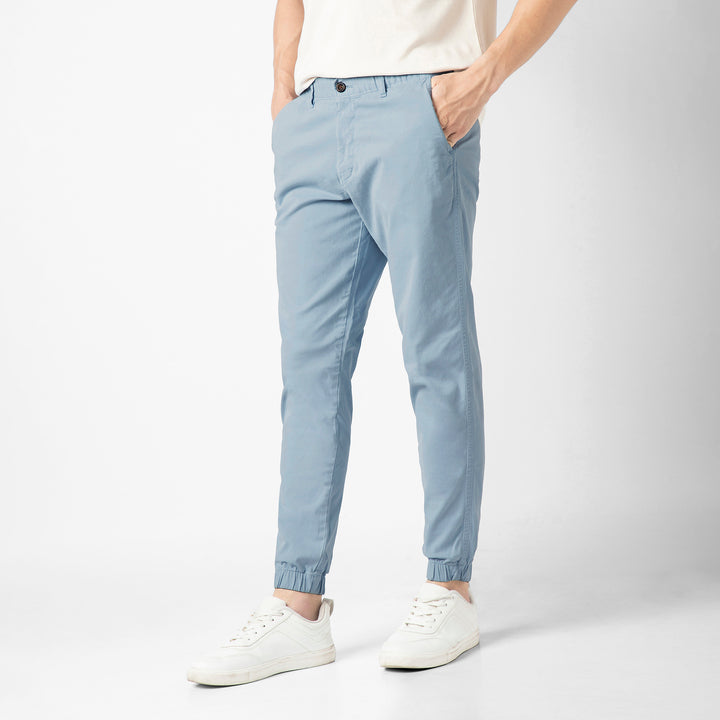 A person stands at an angle in the Coastal light blue Stretch Jogger with one hand tucked into the side pocket, wearing a cream tee and white sneakers against a plain light grey background.