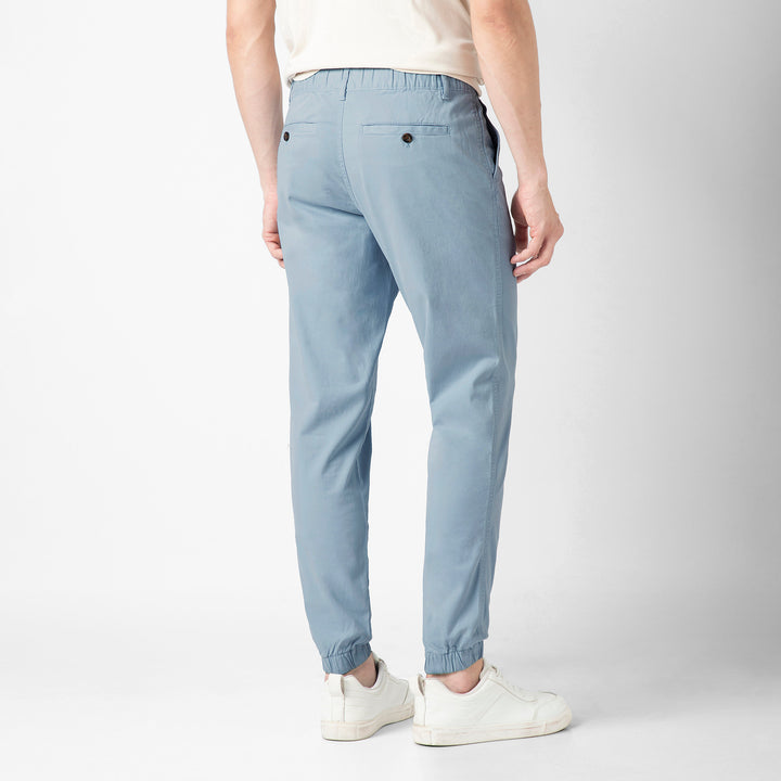 A person stands with their back to the camera in the Coastal light blue Stretch Jogger, showing the buttoned back pocket and elastic ankle cuffs, with a cream tee and white sneakers.