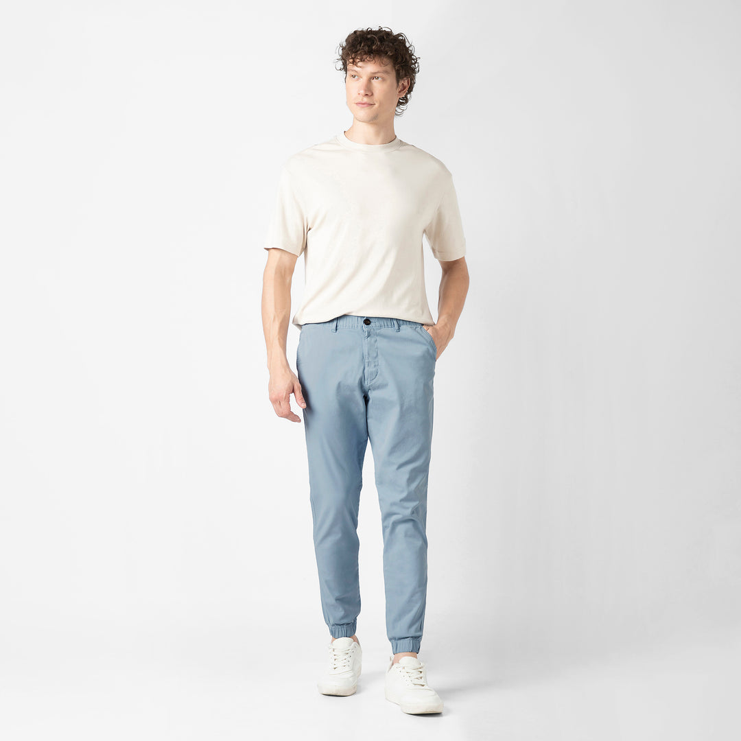 A person stands facing the camera in the Coastal light blue Stretch Jogger with a cream short-sleeve tee and white sneakers, shown head to toe against a plain light grey background.