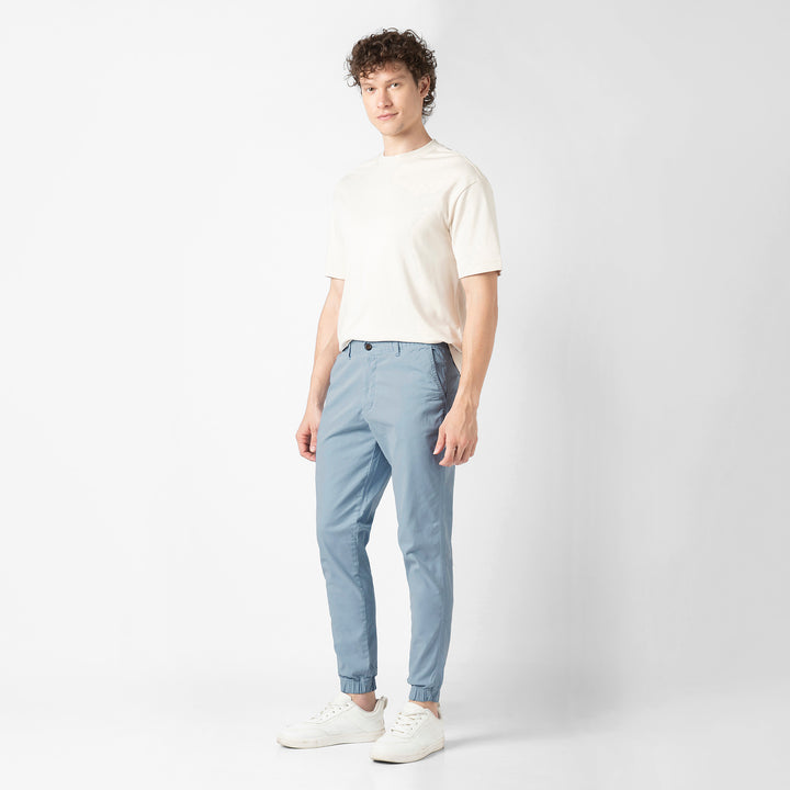A person stands at a three-quarter angle in the Coastal light blue Stretch Jogger with one hand in the pocket, wearing a cream tee and white sneakers, shown head to toe.