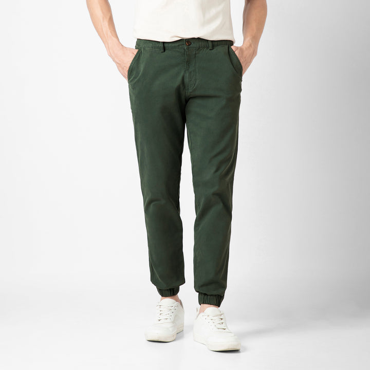 A person wears the Stretch Jogger in Dark Sage with an elastic waistband, paired with a cream short-sleeve tee and white sneakers, against a plain light grey background. The image is cropped at the shoulders.