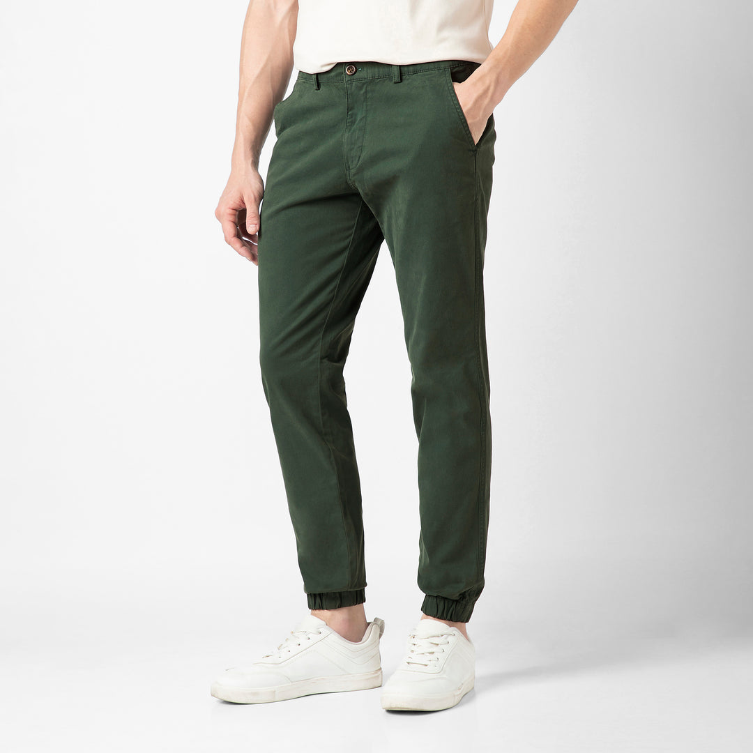 A person stands at an angle in the Dark Sage Stretch Jogger with one hand tucked into the side pocket, wearing a cream tee and white sneakers against a plain light grey background.