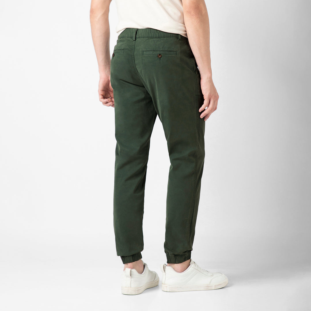 A person stands with their back to the camera in the Dark Sage Stretch Jogger, showing the buttoned back pocket and elastic ankle cuffs, with a cream tee and white sneakers.