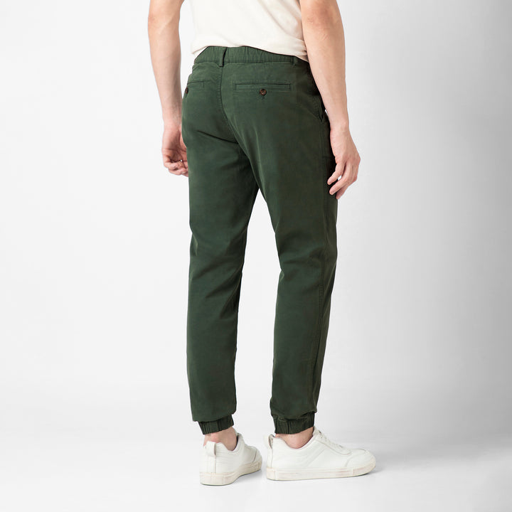 A person stands with their back to the camera in the Dark Sage Stretch Jogger, showing the buttoned back pocket and elastic ankle cuffs, with a cream tee and white sneakers.