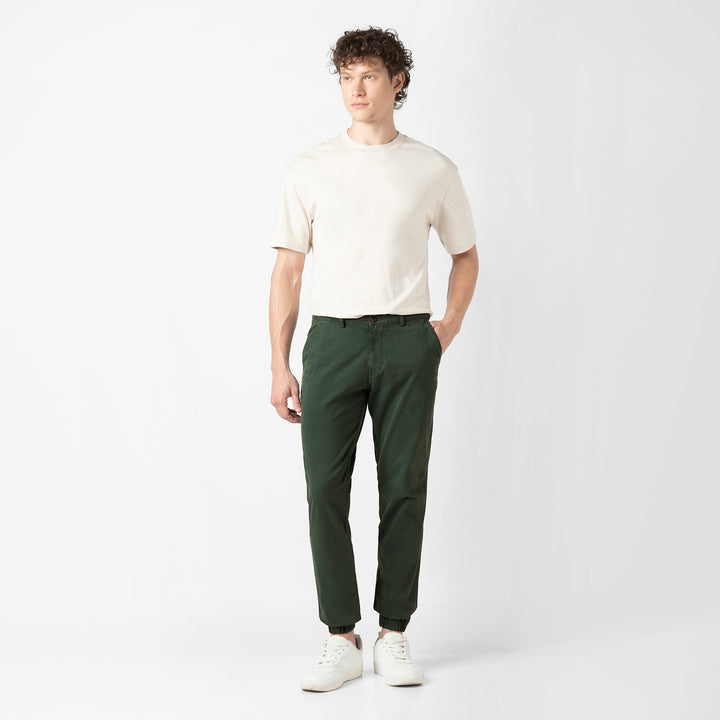 A person stands facing the camera in the Dark Sage Stretch Jogger with a cream short-sleeve tee and white sneakers, shown head to toe against a plain light grey background.