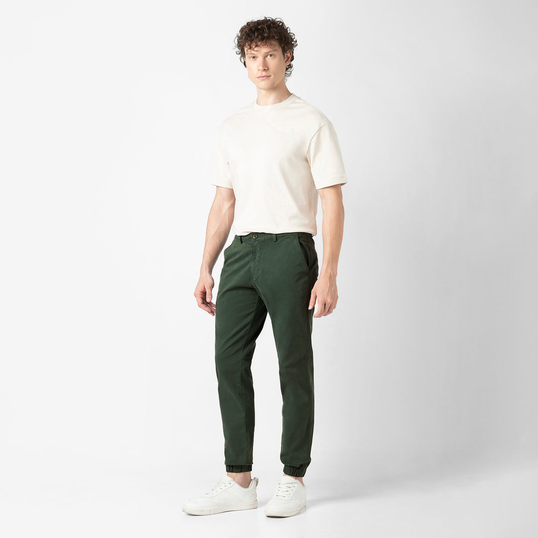 A person stands at a three-quarter angle in the Dark Sage Stretch Jogger with one hand in the pocket, wearing a cream tee and white sneakers, shown head to toe.