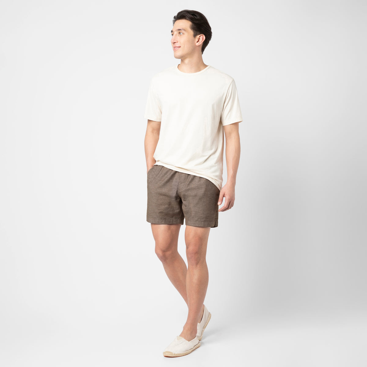A man stands against a white background, wearing a cream T-shirt, brown Stretch Linen Short, and cream slip-on shoes. He faces slightly to the side with one hand in his pocket and has a relaxed expression.