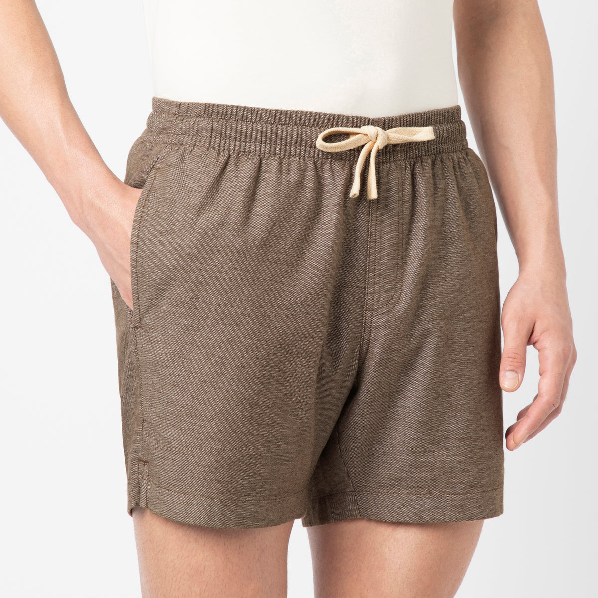 A person wearing a lightweight, relaxed light shirt and the Stretch Linen Short in brown with an elastic waistband stands with one hand in their pocket against a plain background.