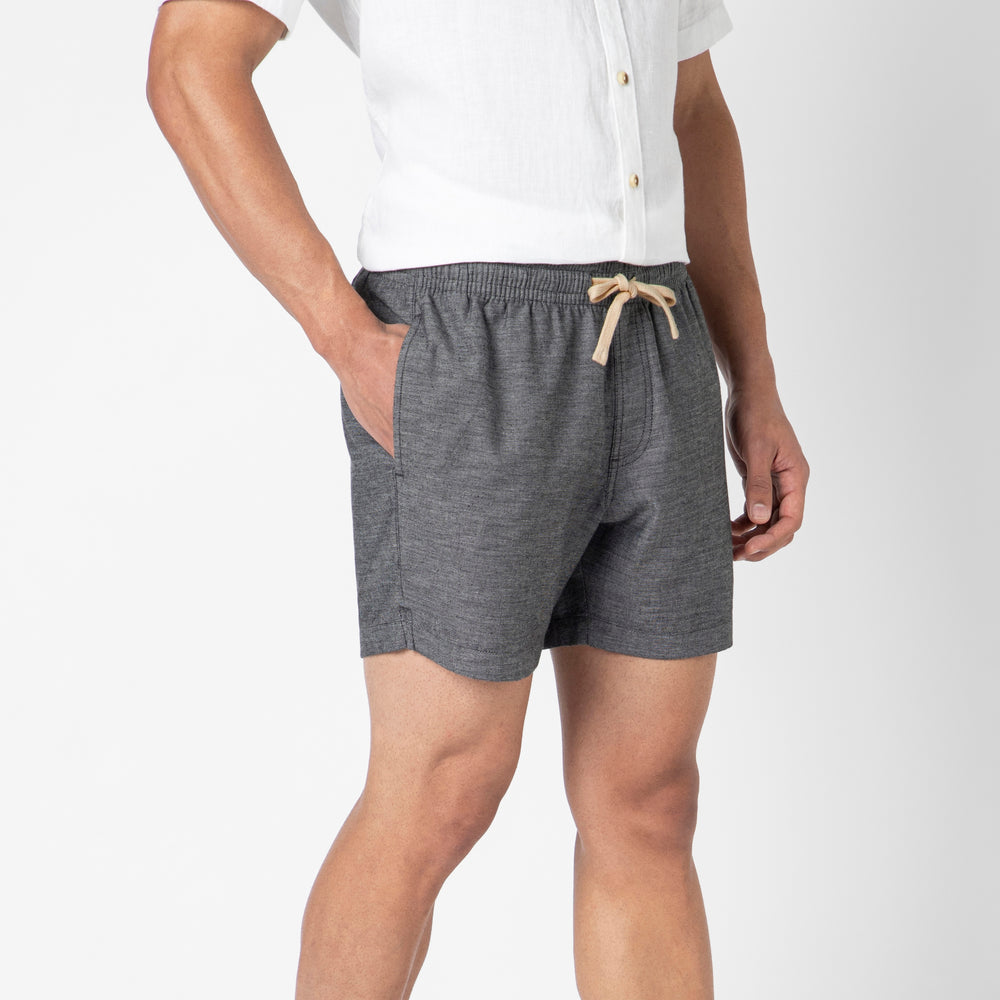 Wearing the Stretch Linen Short in gray and a premium European linen white short-sleeve button-up, a person stands with hands in pockets against a plain white background, highlighting a lightweight, relaxed summer style.