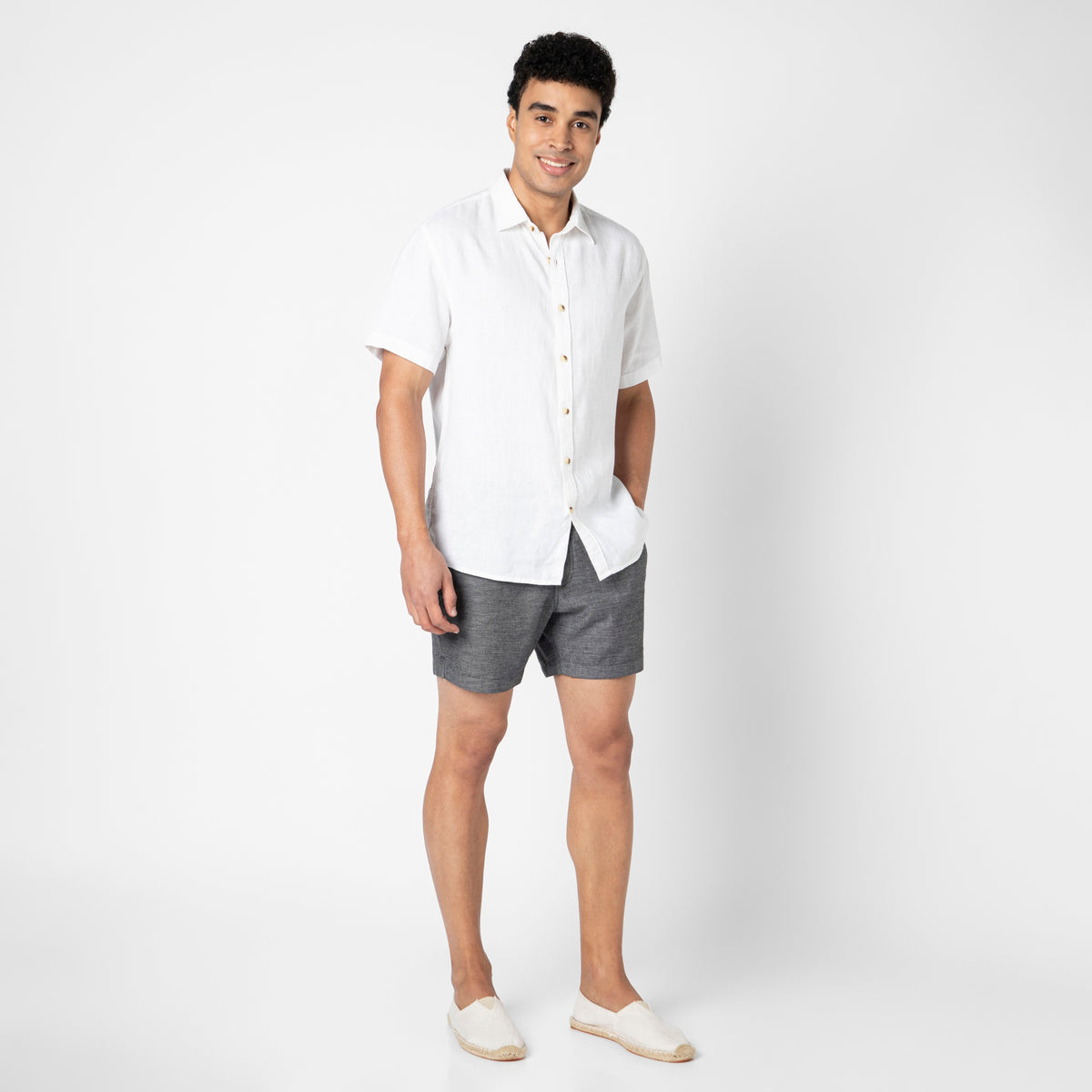 A man stands against a white background wearing a short-sleeved white button-up shirt, gray Stretch Linen Short, and white slip-on shoes, smiling with one hand in his pocket.