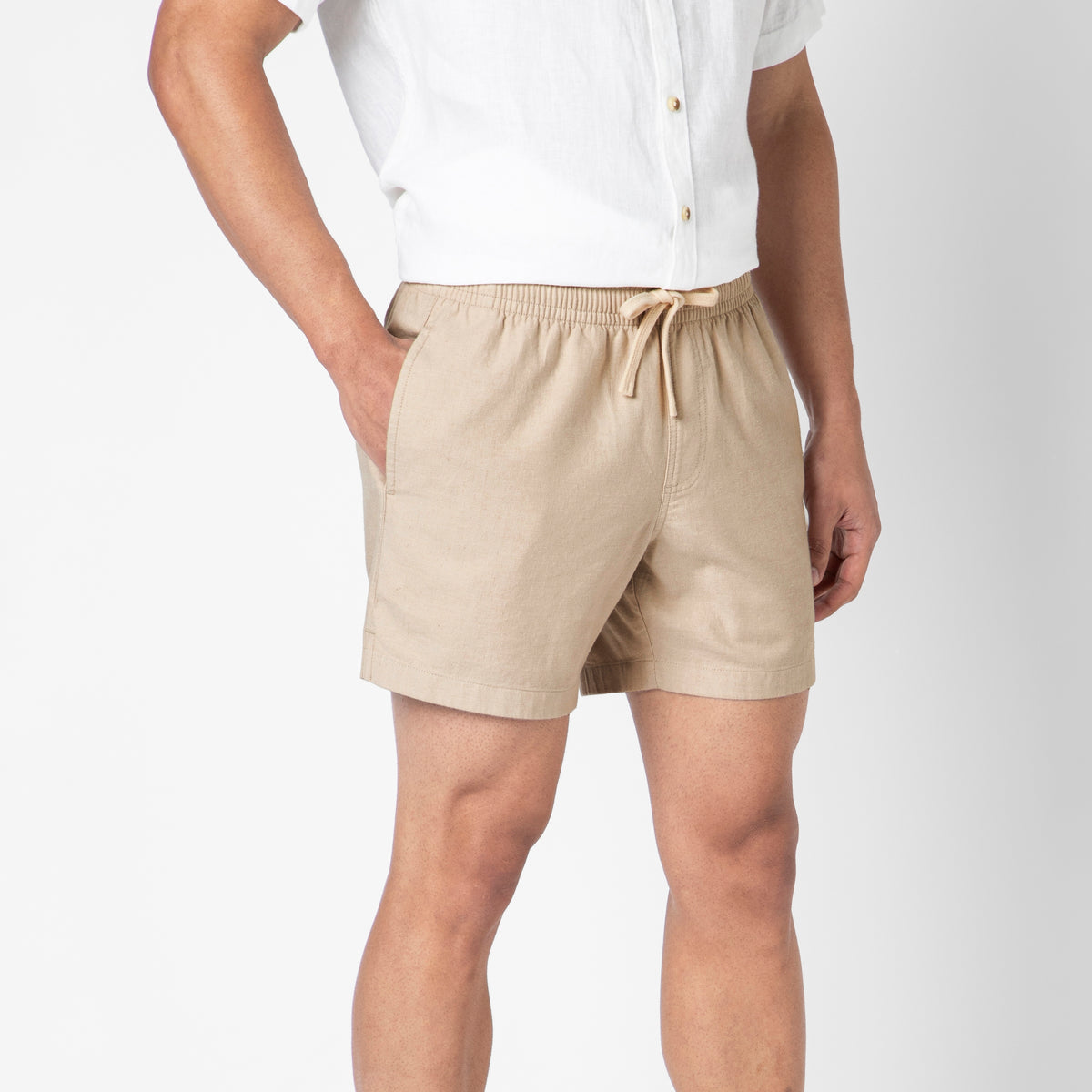 A person wearing beige Stretch Linen Shorts with hands in pockets, paired with a white short-sleeve button-up shirt, stands against a plain white background.