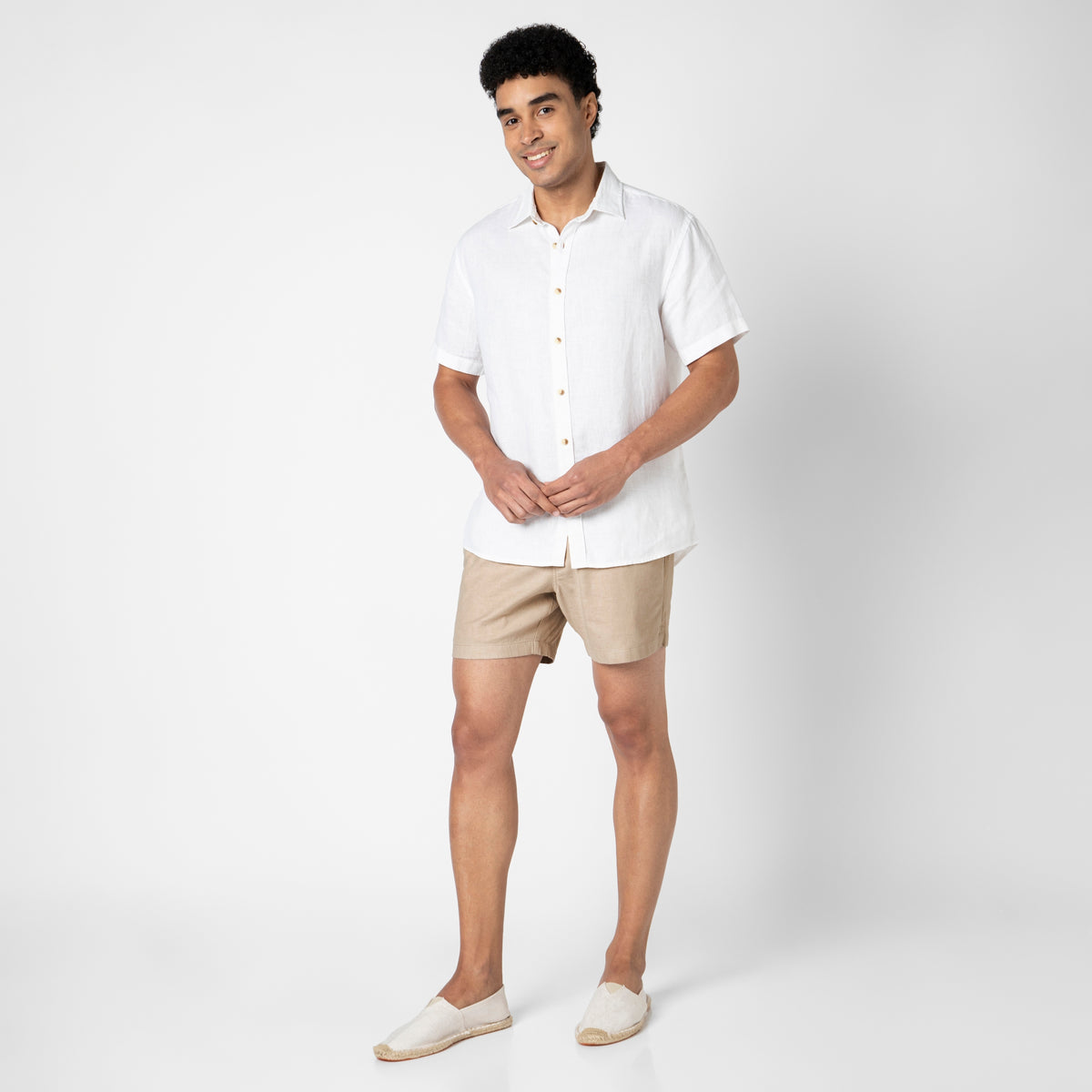 A man smiles against a white background, wearing a short-sleeved white button-up shirt, tan Stretch Linen Short, and white slip-on shoes. His loosely clasped hands highlight the comfort of the lightweight shorts.