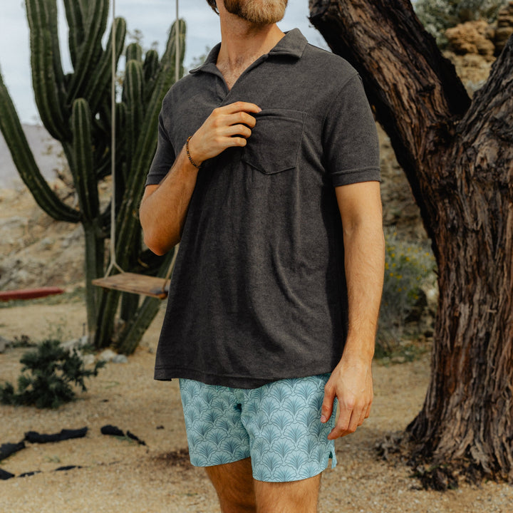 A man wears the Stretch Swim Trunk (Clearance) in Shell, 5.5" inseam, with a dark grey Coastal Terry Polo, standing in front of a cactus.