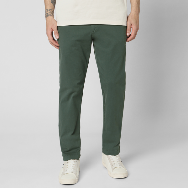 A person wearing Dark Sage Stretch Chino Pant with an elastic waistband, cream shirt, and white sneakers stands against a plain light gray background. Only the lower half of the body is visible.