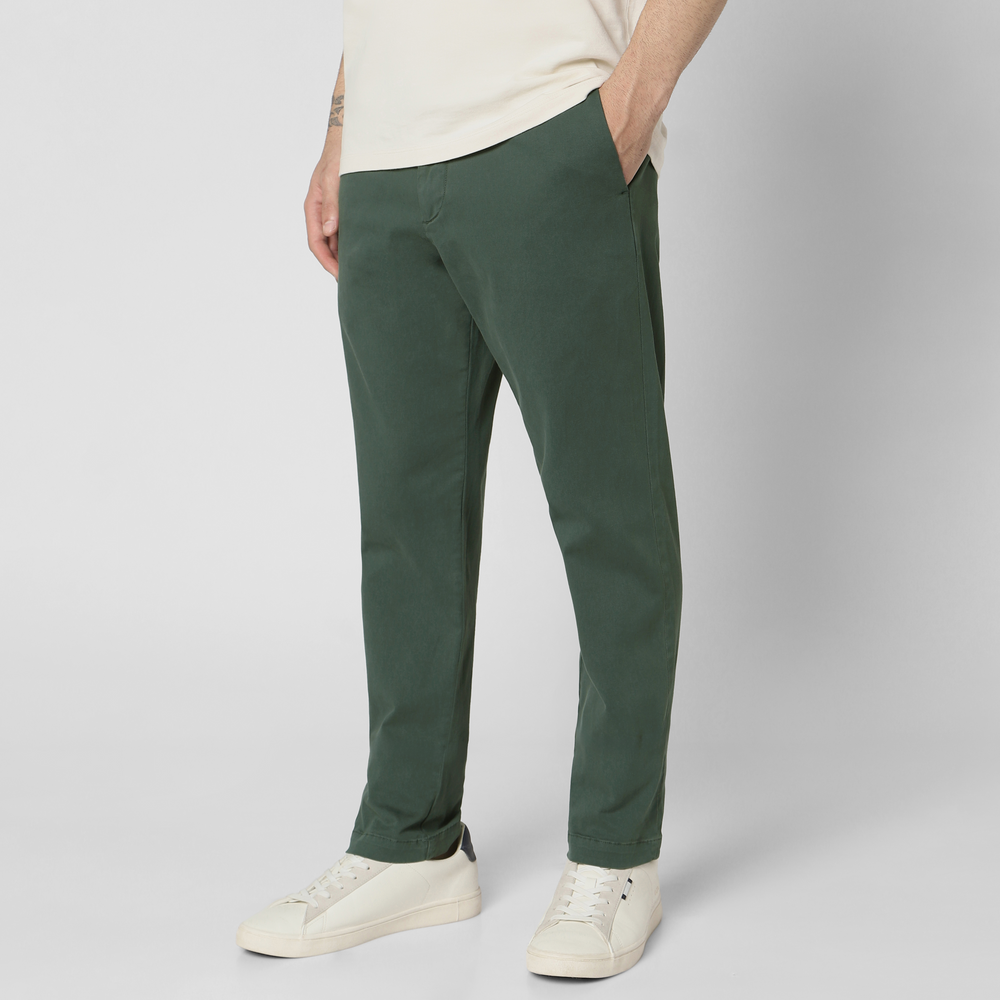 A person wearing dark green Stretch Chino Pant, a cream shirt, and white sneakers stands with hands in pockets against a plain light background. The upper part of the head is not visible.