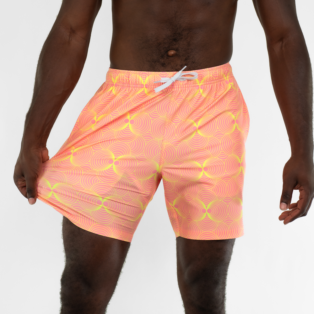 A person wears the Stretch Swim Trunk (Clearance) in peach with a yellow geometric swirl pattern and white drawstring, standing against a plain white background. The water-resistant shorts feature stretch fabric for comfort.