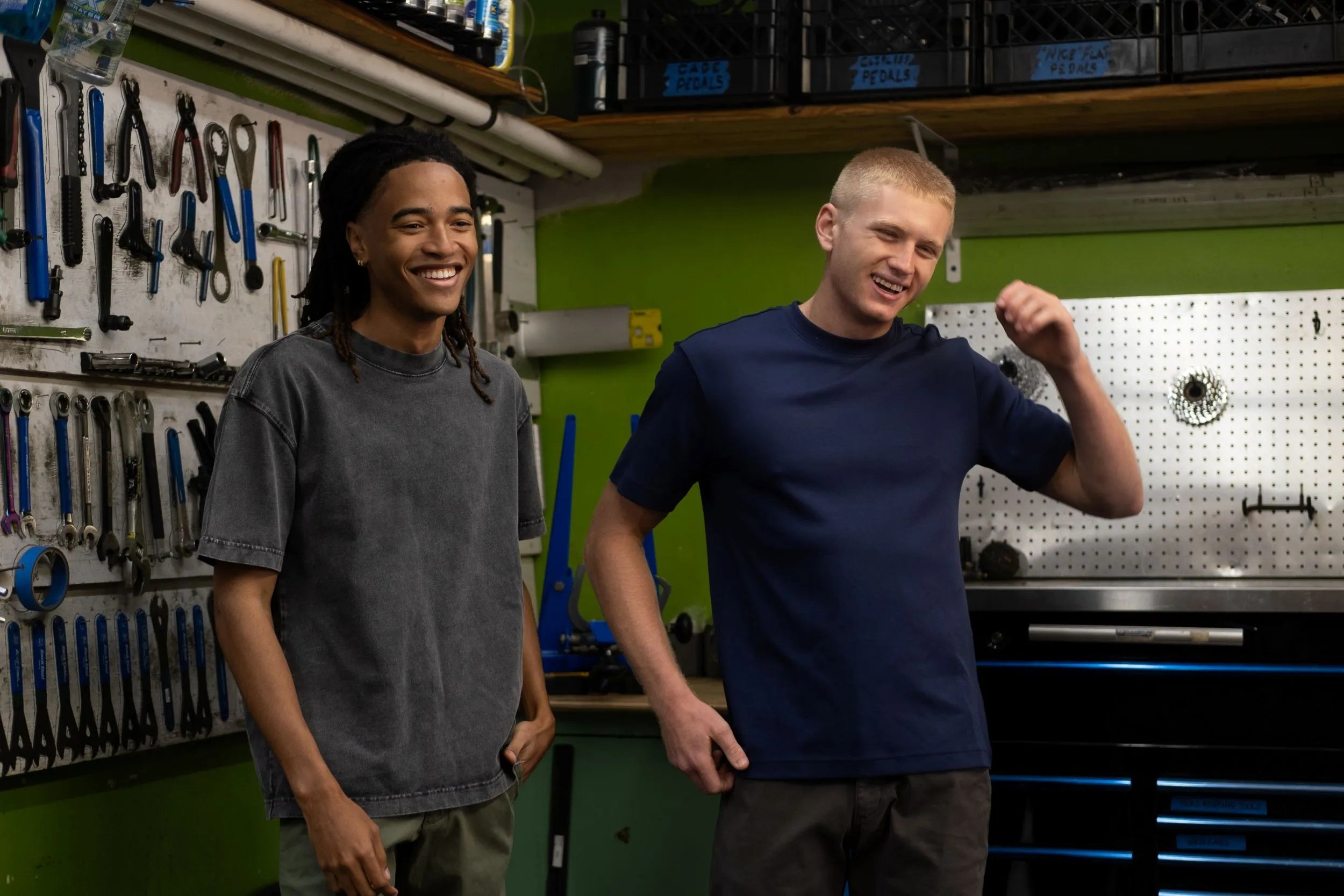 Two men standing in a workshop with tools and equipment in the background. Man on left wearing Boxy Tee in Shadow and Man on right wearing Midweight Supima Tee in Navy. Shop our trending styles. Shop Best Sellers now.