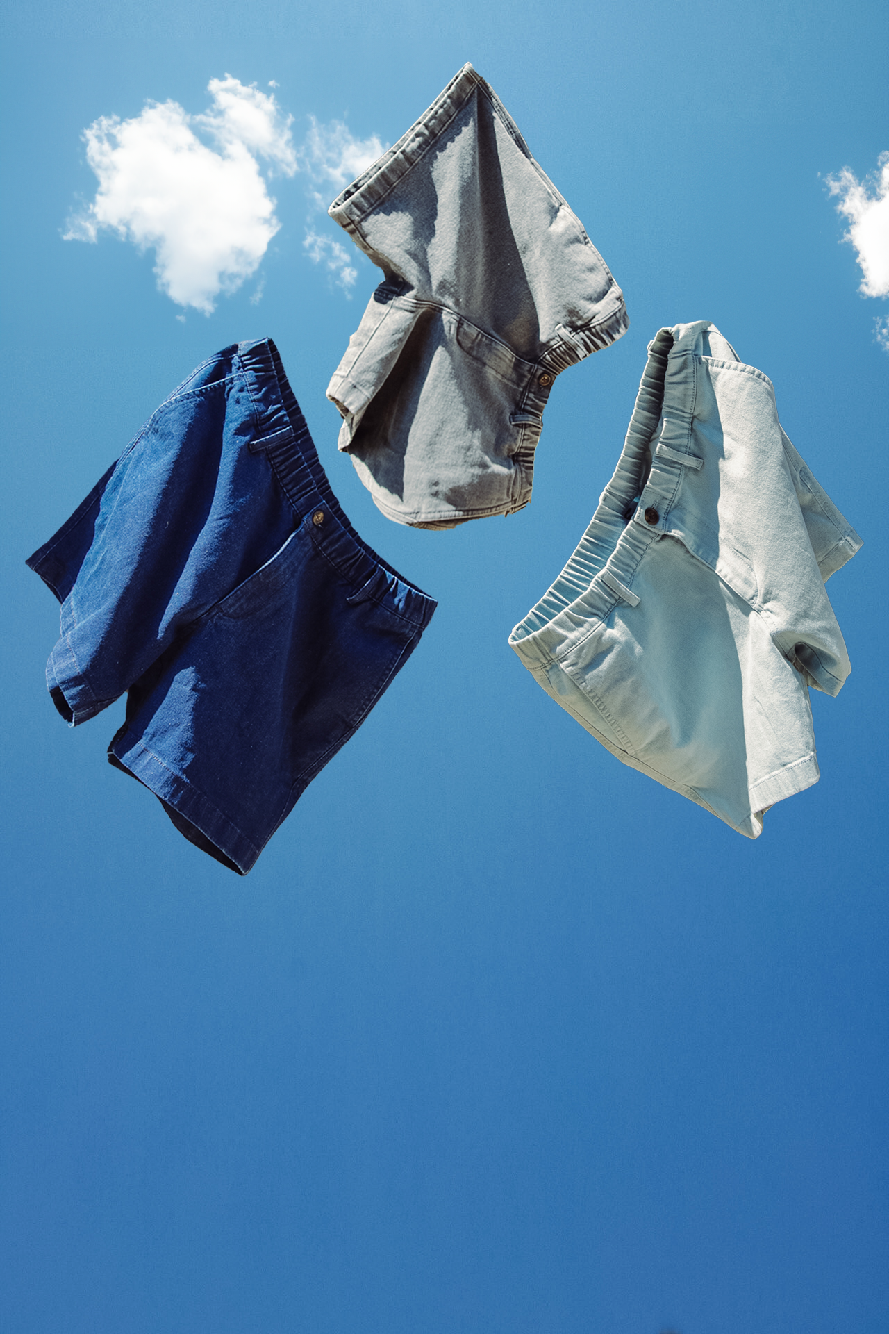 Three pairs of Bearbottom Stretch Denim Short in Dark Wash, Light Wash, and Black, suspended mid-air against a blue sky